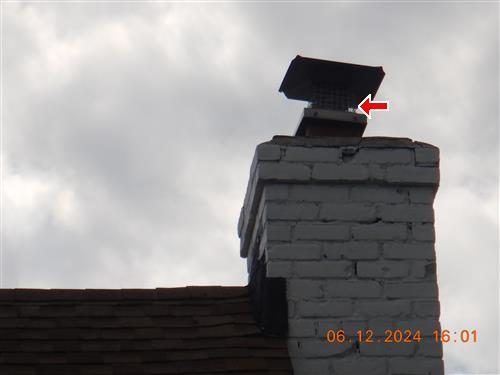White-painted brick chimney against a cloudy sky with a metal rain cap on top; a red arrow points at the cap.