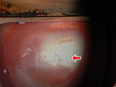Close-up inside a boiler showing a reddish firebox surface covered in a network of fine cracks, with a red arrow pointing at them.