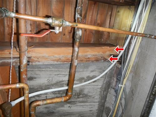 Basement ceiling with copper pipes and a shut-off valve in front of wood framing, two red arrows pointing at the joists on the right.