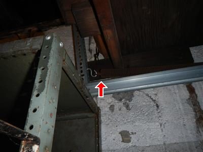Garage door lift spring and track beside a metal shelf, with a red arrow pointing up at the spring.