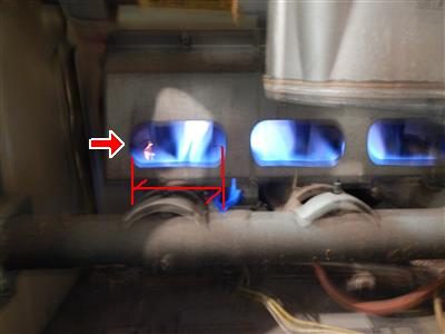 Blue gas burner flames inside a furnace, the left flame spreading wider than the others, marked with a red arrow and bracket.