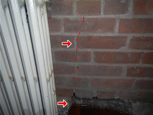 Interior brick wall beside a white radiator with a vertical crack traced in red dashes and two red arrows.