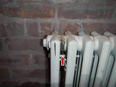 White cast iron radiator against a brick wall with a dark stain at the end section, red arrow.