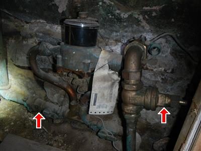 Brass water meter and shut-off valves on pipes in a dark basement with a paper tag, two red arrows.
