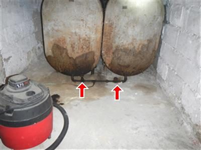 Two rusted oil tanks in a block-walled room with red arrows pointing to the shut-off valves at their base.