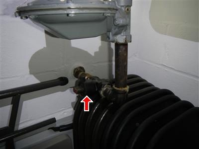 Gas shut-off valve on a pipe beside a cast-iron boiler in a basement, marked with a red arrow.