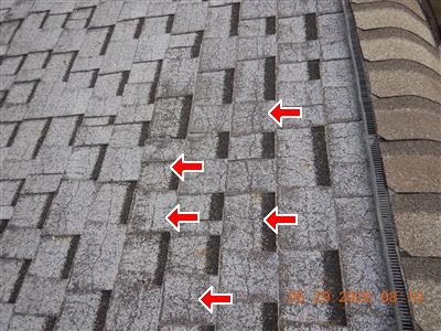 Grey asphalt shingle roof near the ridge with five red arrows pointing at cracked shingles.