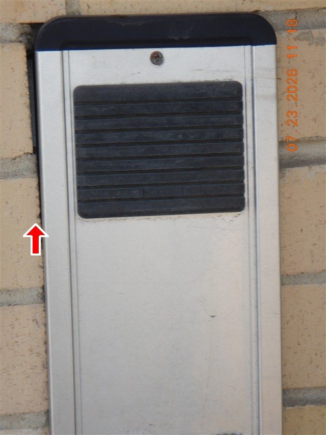 Metal intercom panel set into a tan brick wall with a visible gap along its left edge; a red arrow points at the gap.