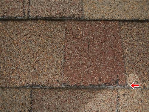 Close-up of worn asphalt shingles with a red arrow at a corner where the fibreglass mat is exposed.