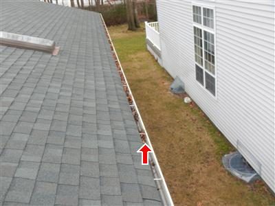 Roof edge gutter full of leaves beside a house wall, with a red arrow pointing into the gutter.