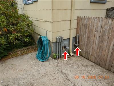 Oil fill and vent pipes at the base of a house wall beside a wood fence, marked with two red arrows.