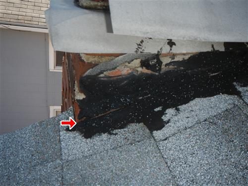 Base of a brick chimney on an asphalt shingle roof with black tar flashing; a red arrow points at the flashing edge.