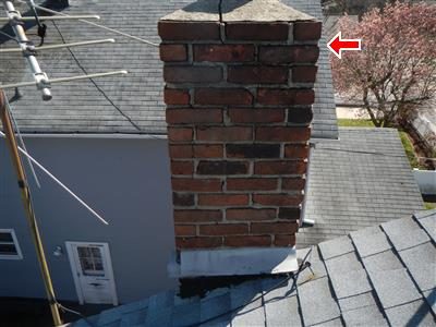Top of a brick chimney with a concrete cap and eroded mortar joints, red arrow at the cap, seen from the roof.