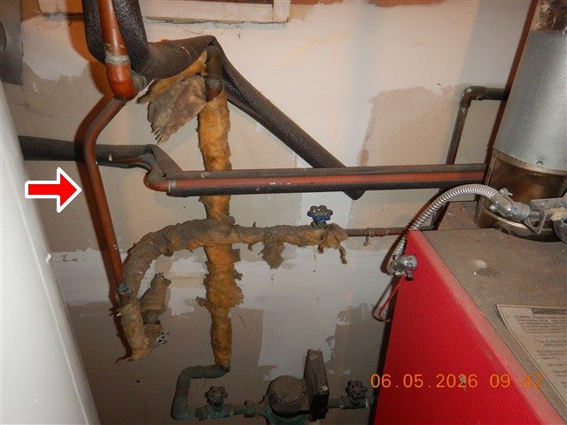 Boiler room with bare copper pipes and old insulated pipes above a red boiler, a red arrow pointing at an uninsulated copper pipe.