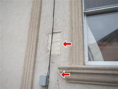 Stucco wall beside a window with a rectangular patch and a cable running down it, two red arrows pointing at the patch and a lower ledge.
