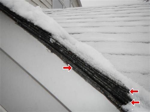 Snow-covered shingle roof edge showing three stacked shingle layers, marked with three red arrows.