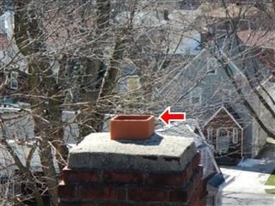 Brick chimney top with a concrete cap and an open clay flue, red arrow pointing at the flue, bare trees behind.