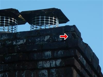 Top of a brick chimney with two metal caps, eroded brick joints and a red arrow at the cracked cement cap.