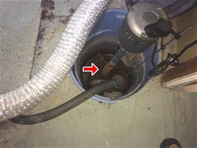 Sump pump sitting in a blue pit in a basement floor with flexible duct beside it, a red arrow pointing at the pump shaft.