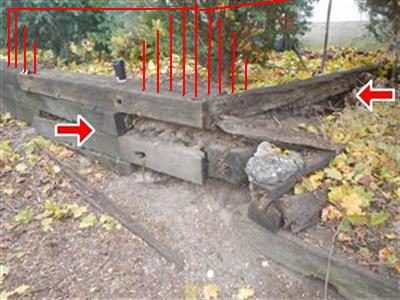 Wood railroad tie retaining wall with split, crumbling timbers, marked with red arrows and red hatching.