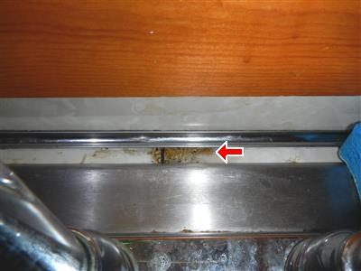 Kitchen counter edge behind a stainless faucet with a dark rotted patch, marked by a red arrow.