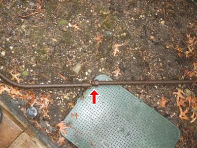 Rusted metal conduit lying across soil in a flower bed beside a green utility cover, marked with a red arrow.