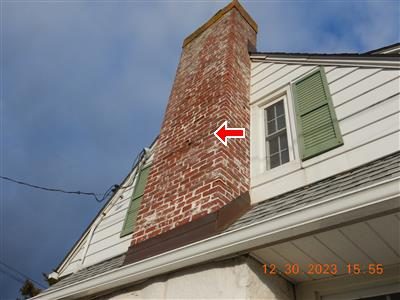 Tall brick chimney against a blue sky with spalled brick faces midway up, marked with a red arrow.