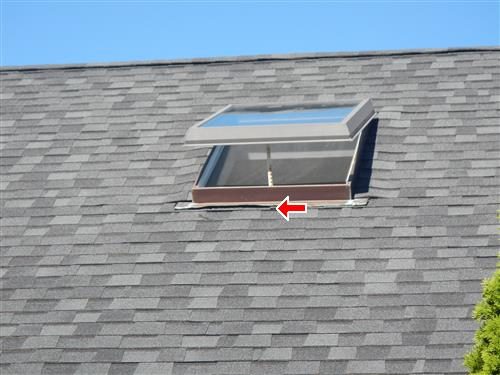 Open skylight on a grey shingle roof with a red arrow pointing at the lower edge of its flashing.