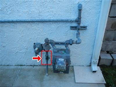 Gas meter and piping on a white stucco exterior wall, with a red box and arrow around the shut off valve.