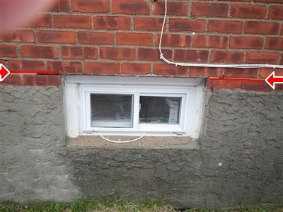 Small basement window set in a brick and stucco wall with red arrows at the steel lintel line above it.