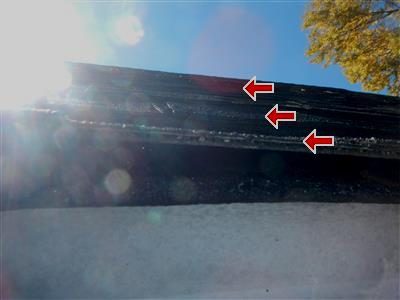 Edge of a lower roof seen from below against bright sun, three red arrows marking the stacked shingle layers.