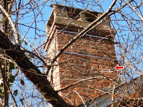 Tall brick chimney seen through bare tree branches against a blue sky, with a red arrow pointing at its right side.