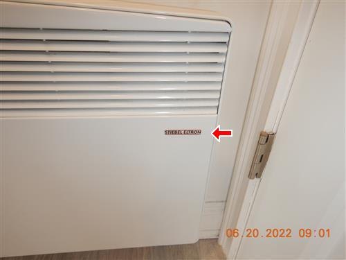 A wall-mounted electric convector heater beside a door frame, with an arrow at its data label.