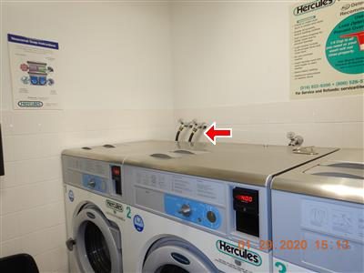 Commercial washing machines in a laundry room with a red arrow pointing to the supply hoses behind them.