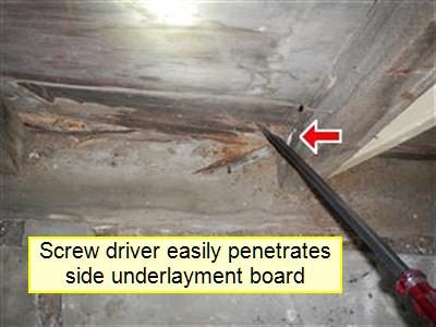 Screwdriver tip pressed into a deteriorated wood underlayment board at the base of a wall, marked by a red arrow.