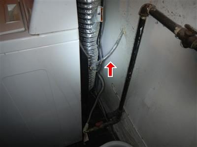 Exposed armoured BX cable running up the wall behind a clothes dryer, marked with a red arrow.