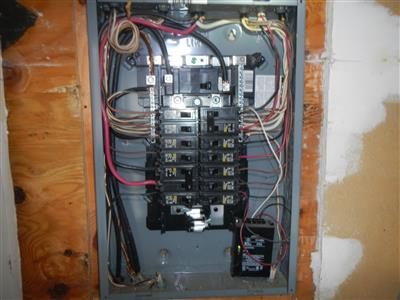 Open grey electrical panel with rows of breakers and wiring, mounted on plywood in a garage.