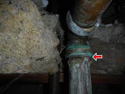 Corroded green pipe joint seen through a ceiling hatch below a bathtub, marked by a red arrow.