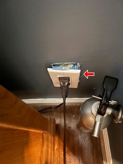 Red arrow at an outlet cover pulled away from a damaged wall opening, cord plugged in