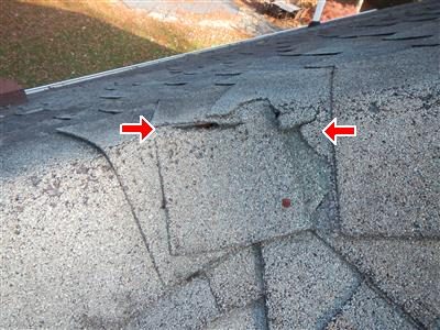 Asphalt ridge capping at a roof peak corner with a visible crack, marked by two red arrows.