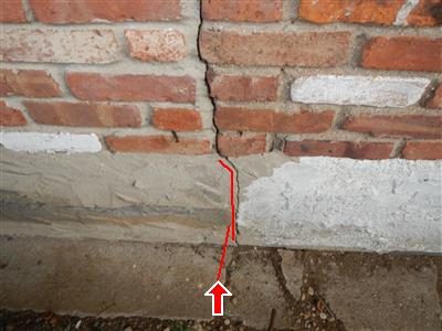 Brick and parged foundation wall with a red line tracing a vertical crack and a red arrow at its base.