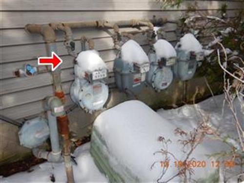 A bank of four gas meters mounted on house siding under snow, one arrowed.