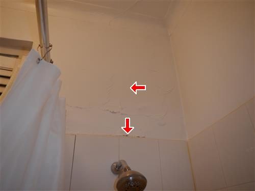 Bathroom wall above a shower head with flaking plaster and a stained seam at the tile line, marked by red arrows.