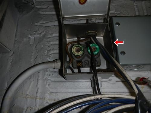 Small metal electrical box with two screw-in fuses and wiring, mounted on a painted masonry wall, marked by a red arrow.