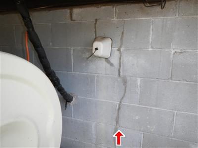 Grey concrete block foundation wall with a vertical crack running top to bottom, marked by a red arrow at the base.