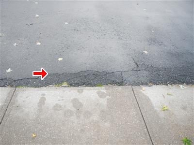 Asphalt driveway edge meeting a concrete sidewalk with a line of cracks, marked by a red arrow.