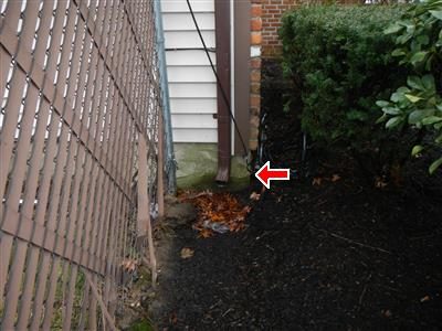 Leader pipe elbow at the base of a house wall by a chain-link fence, red arrow at the elbow discharging onto soil.