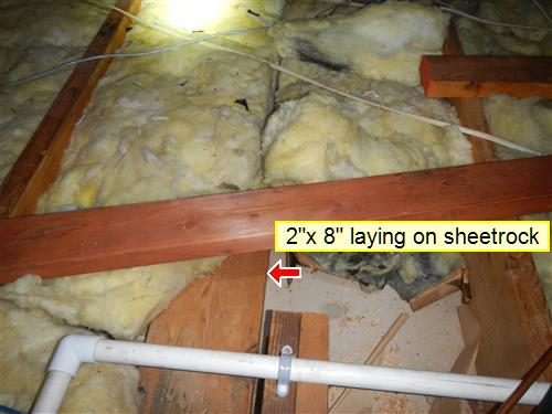Attic with fibreglass insulation between joists and a loose 2x8 lying on the sheetrock, marked with a red arrow.