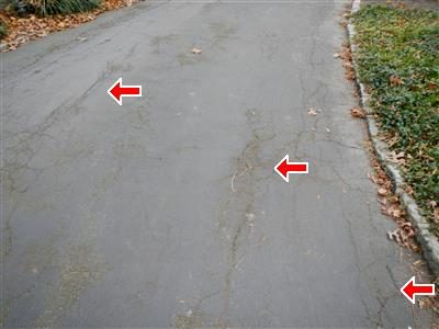 Asphalt driveway with several cracks marked by three red arrows, grass and leaves along the edge.