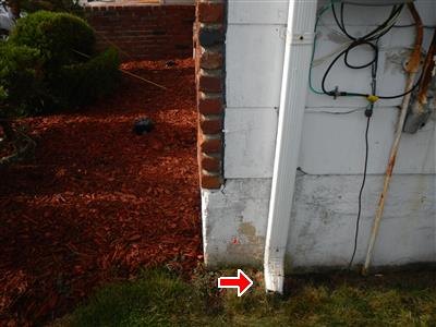A downspout terminating at a foundation corner near grade, with an arrow at the discharge point.
